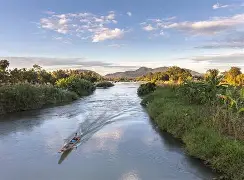 Long narrow boat traveling along a calm river surrounded by lush green vegetation and distant mountains under soft sunlight