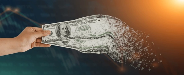 Hand holding a one‑hundred‑dollar bill disintegrating into particles against a dark financial chart background, symbolizing money loss and economic pressure.