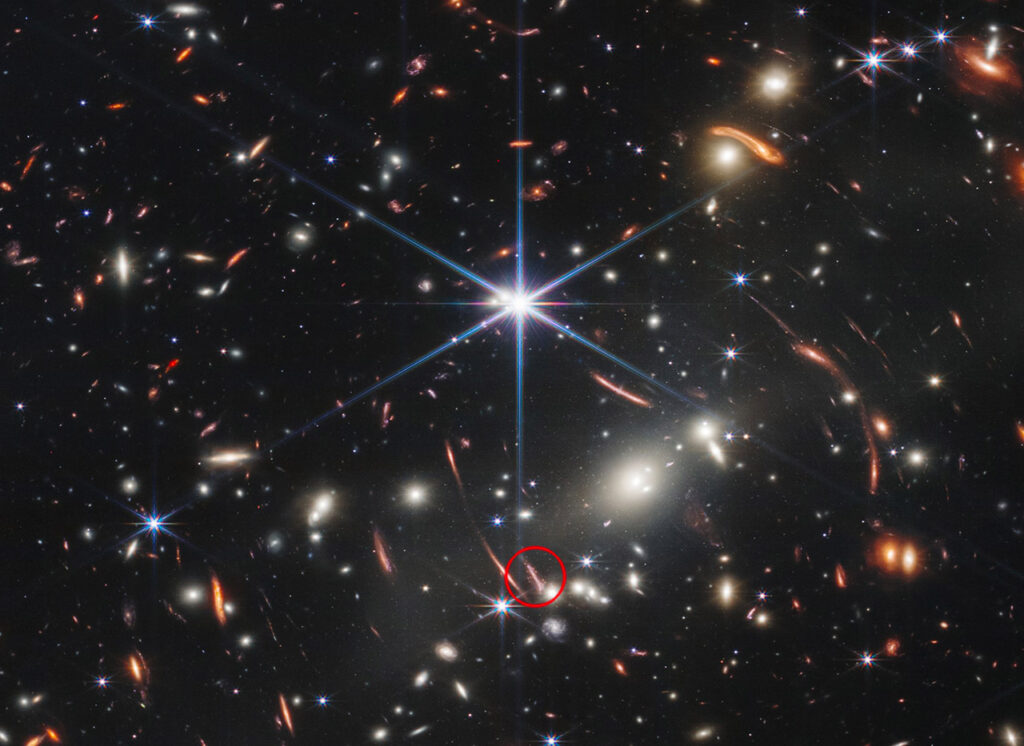 A deep‑field James Webb Space Telescope image showing dozens of distant galaxies scattered across space, with a faint, reddish ancient galaxy highlighted near the center as a potential dark‑star candidate.
