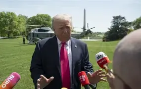 A political figure speaking to reporters outdoors near the Washington Monument, with microphones from major news outlets and a helicopter in the background