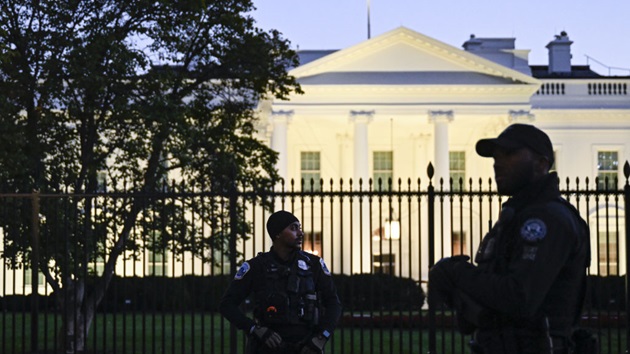 Security officers standing guard in front of the White House behind a tall metal fence during evening hours