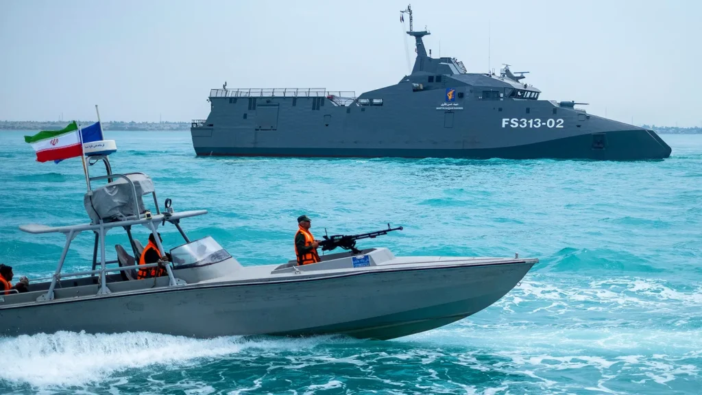 “Iranian patrol boat with armed crew and national flag operating near a large military warship during naval activity in the Strait of Hormuz