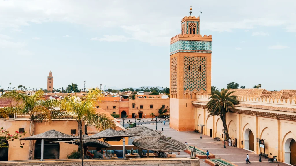 Marrakech and the High Atlas – view of The Medina with its traditional mosque, ornate minaret, palm trees, and sunlit Moroccan architecture.