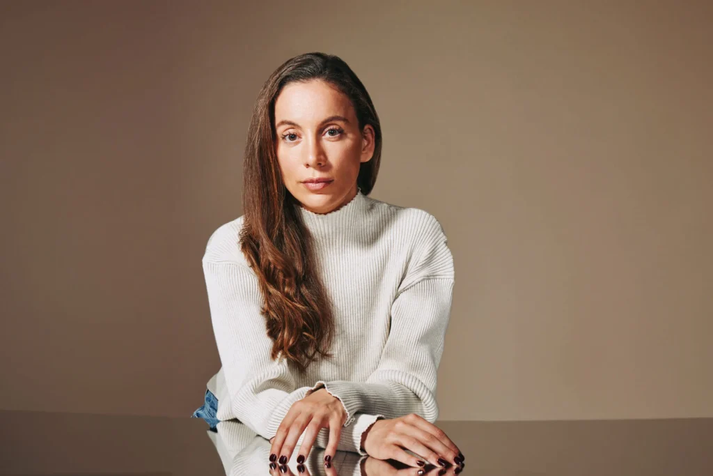 Woman sitting at a reflective glass table in soft neutral lighting A woman wearing a light ribbed sweater sits at a reflective glass surface, her hands resting gently on the table, with soft lighting highlighting the texture of the fabric and her hair falling over one shoulder.